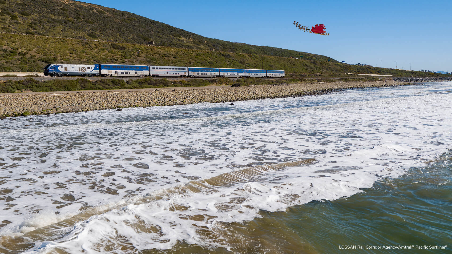 A wide shot of a Pacific Surfliner train from the ocean. Santa’s red sleigh is flying across the blue sky above the tracks.