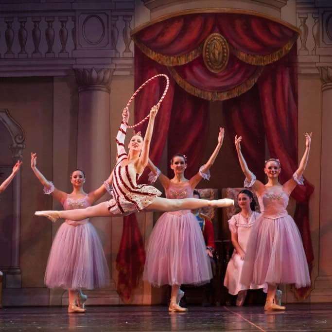 A ballerina holding a hoop does a split leap in front of a group of other ballerinas in pink dresses.